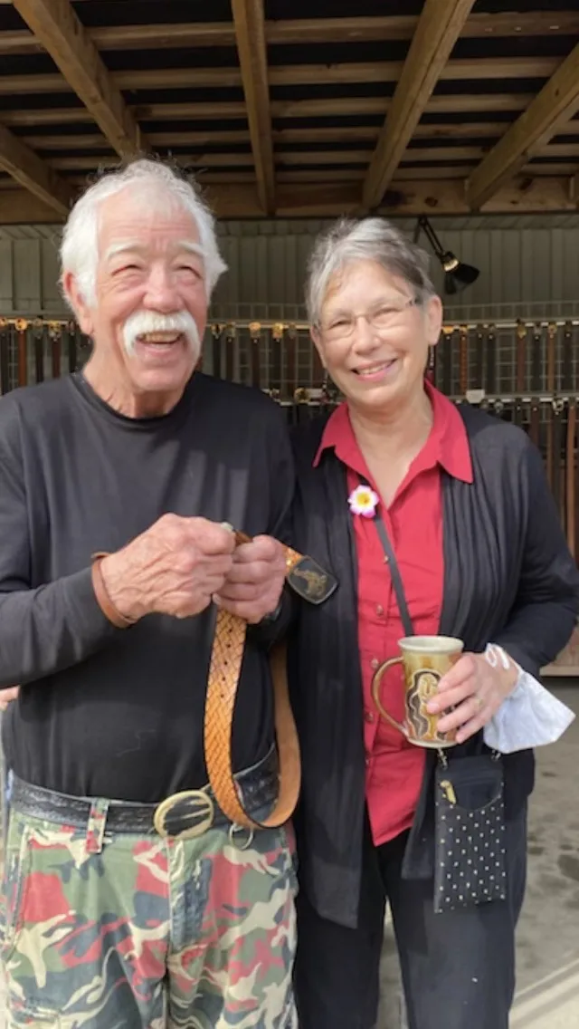 Max and Kathy Whitley, smiling; Max holds a tooled belt, Kathy holds a mug