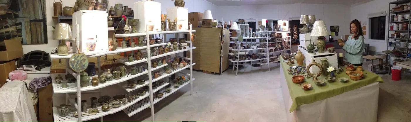 Wide view of long shelves full of pottery, with Kathy arranging a table
